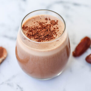 A glass of chocolate smoothie from The New Year Reset Recipe Bundle sits on a marble surface, topped with cocoa powder, with a wooden spoon of cocoa and two dates nearby.