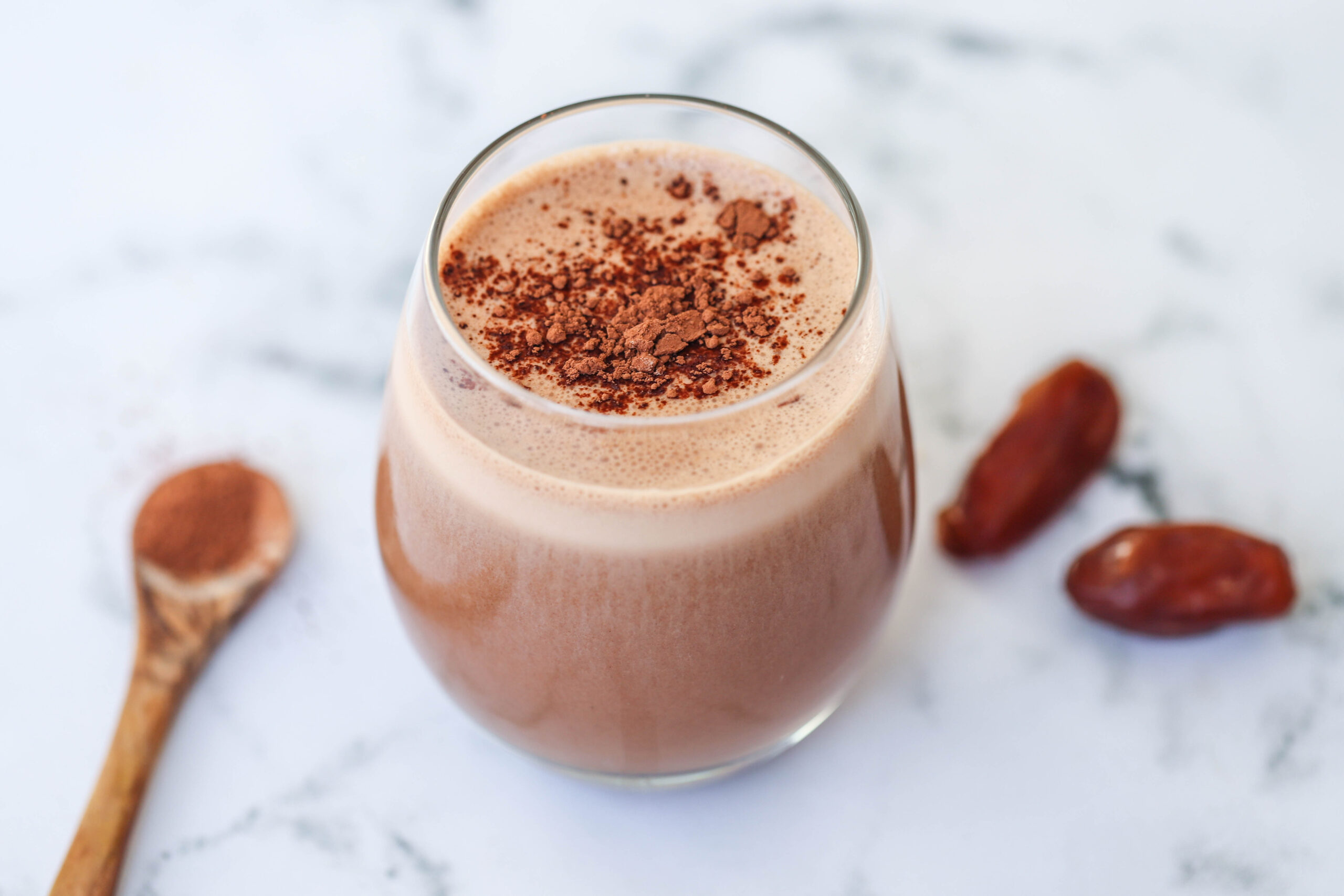 A glass of chocolate smoothie from The New Year Reset Recipe Bundle sits on a marble surface, topped with cocoa powder, with a wooden spoon of cocoa and two dates nearby.