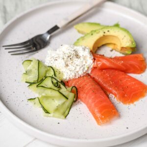 A plate from The New Year Reset Recipe Bundle features smoked salmon, cucumber ribbons, avocado slices, and cottage cheese garnished with herbs. A fork rests on the plate.