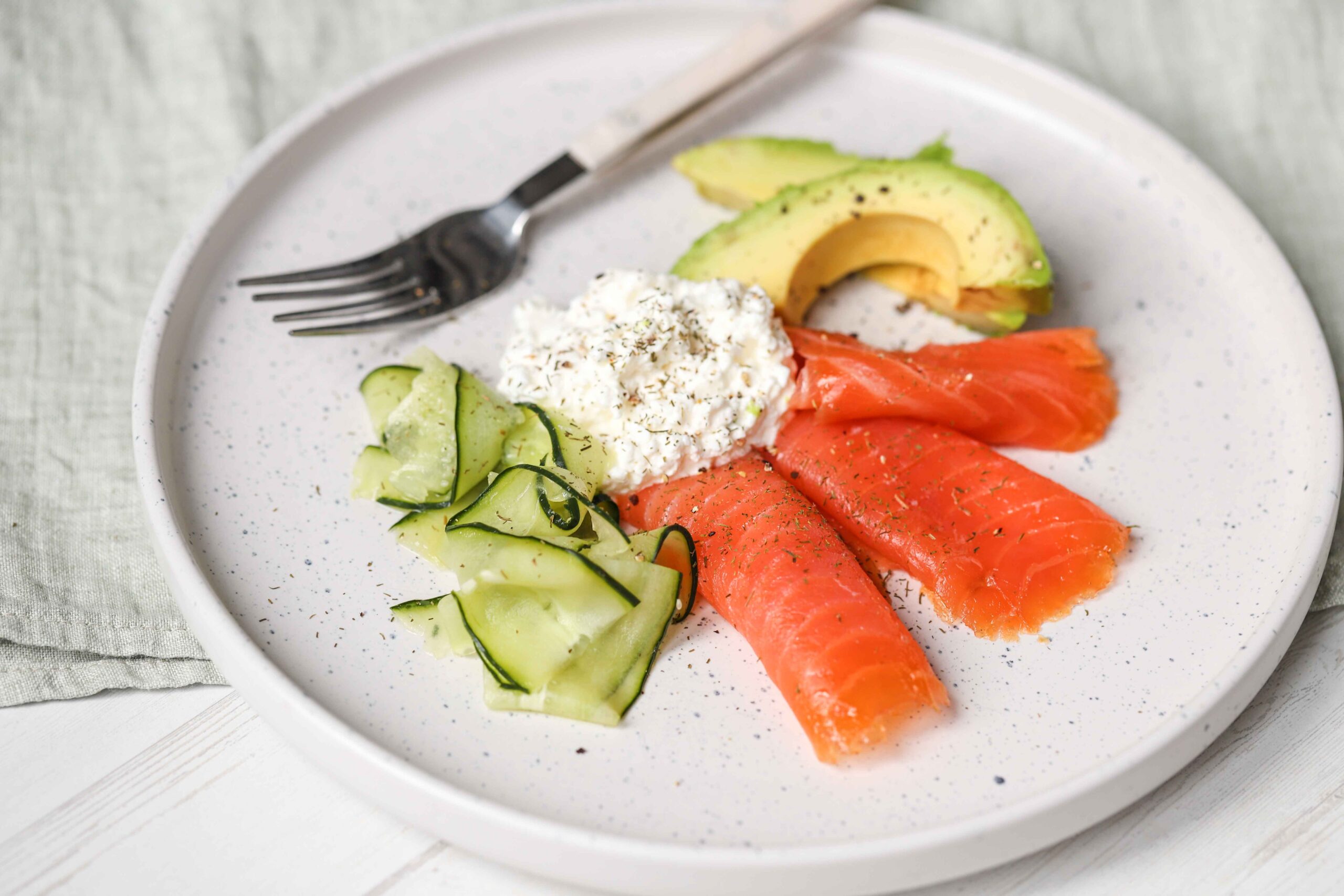 A plate from The New Year Reset Recipe Bundle features smoked salmon, cucumber ribbons, avocado slices, and cottage cheese garnished with herbs. A fork rests on the plate.