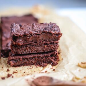 A stack of rich chocolate brownies from The New Year Reset Recipe Bundle sits on parchment paper, with extra brownie pieces, a walnut in the background, and a wooden spoon of cocoa powder in the foreground.