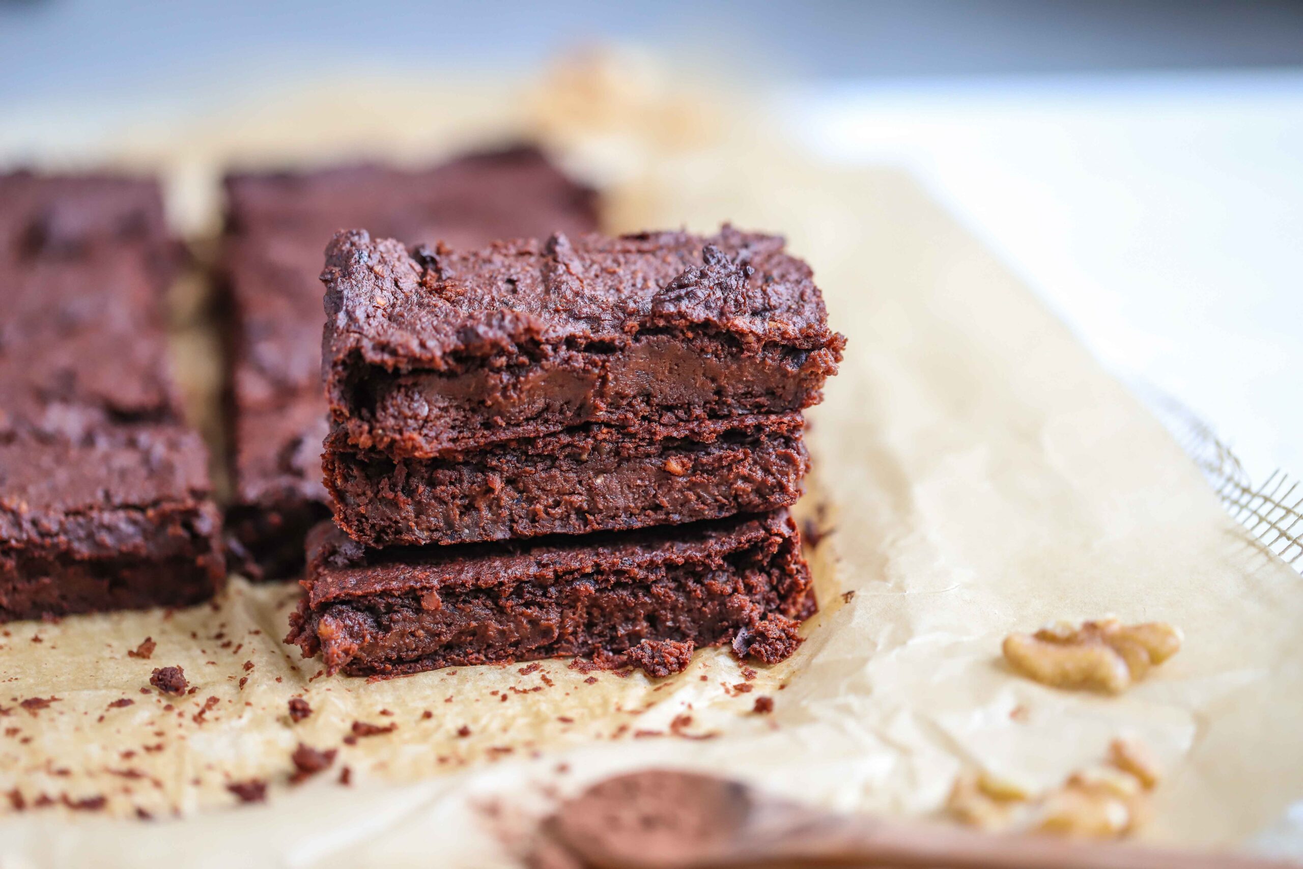 A stack of rich chocolate brownies from The New Year Reset Recipe Bundle sits on parchment paper, with extra brownie pieces, a walnut in the background, and a wooden spoon of cocoa powder in the foreground.