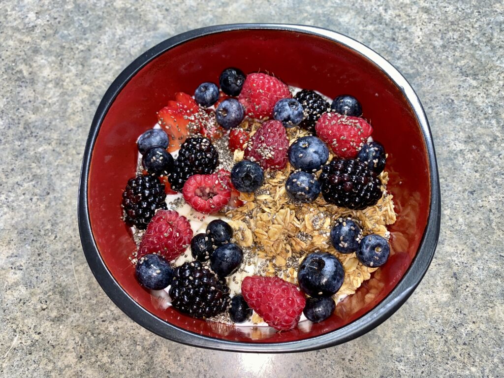 A red and black bowl filled with granola, blueberries, blackberries, raspberries, sliced strawberries, and chia seeds sits on a gray countertop.